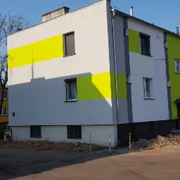 Odnowiona elewacja budynku wielorodzinnego z poziomymi pasami w kolorach żółtym i zielonym, widoczny kontener na gruz i zaparkowane busy, sugerujące prace budowlane w toku.