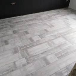 Ułożona podłoga z płytek imitujących drewno w odcieniach szarości, widoczna czarna ściana z gniazdkami elektrycznymi.