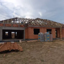 Wznoszenie domu z czerwonej cegły ceramicznej, widoczne drewniane elementy konstrukcji dachu, palety z materiałami budowlanymi oraz sterta desek na pierwszym planie.