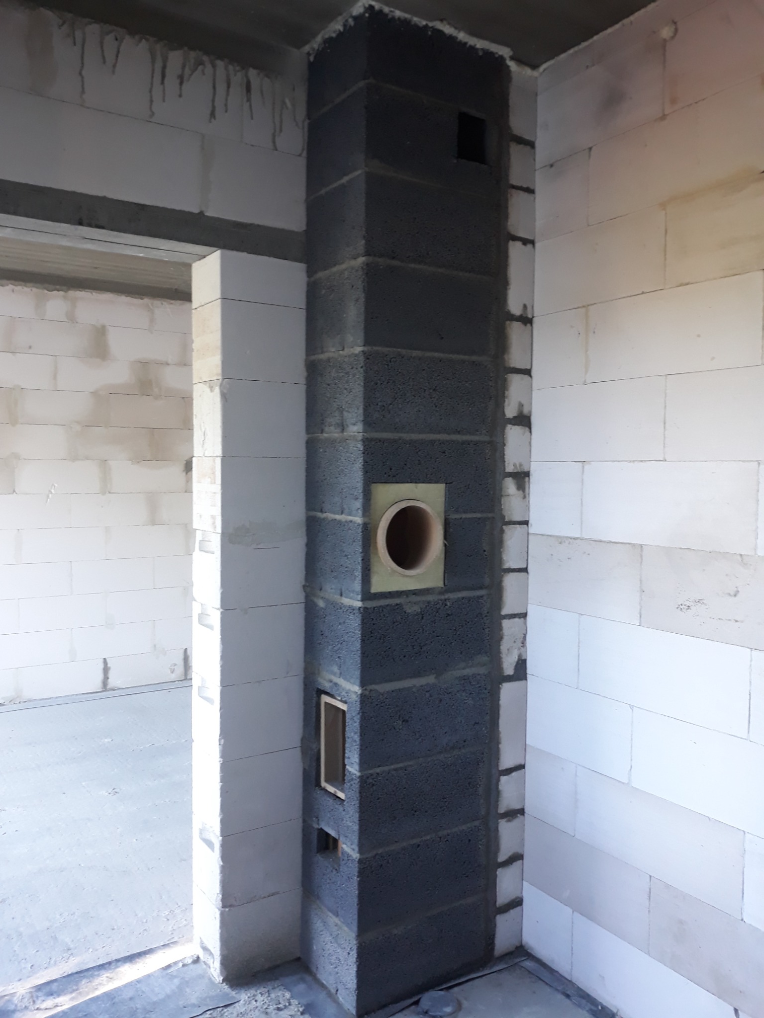 Wnętrze w trakcie budowy, widoczny komin z szarych bloczków z otworem na rurę odprowadzającą spaliny, ściany z białych bloczków, surowy betonowy strop.