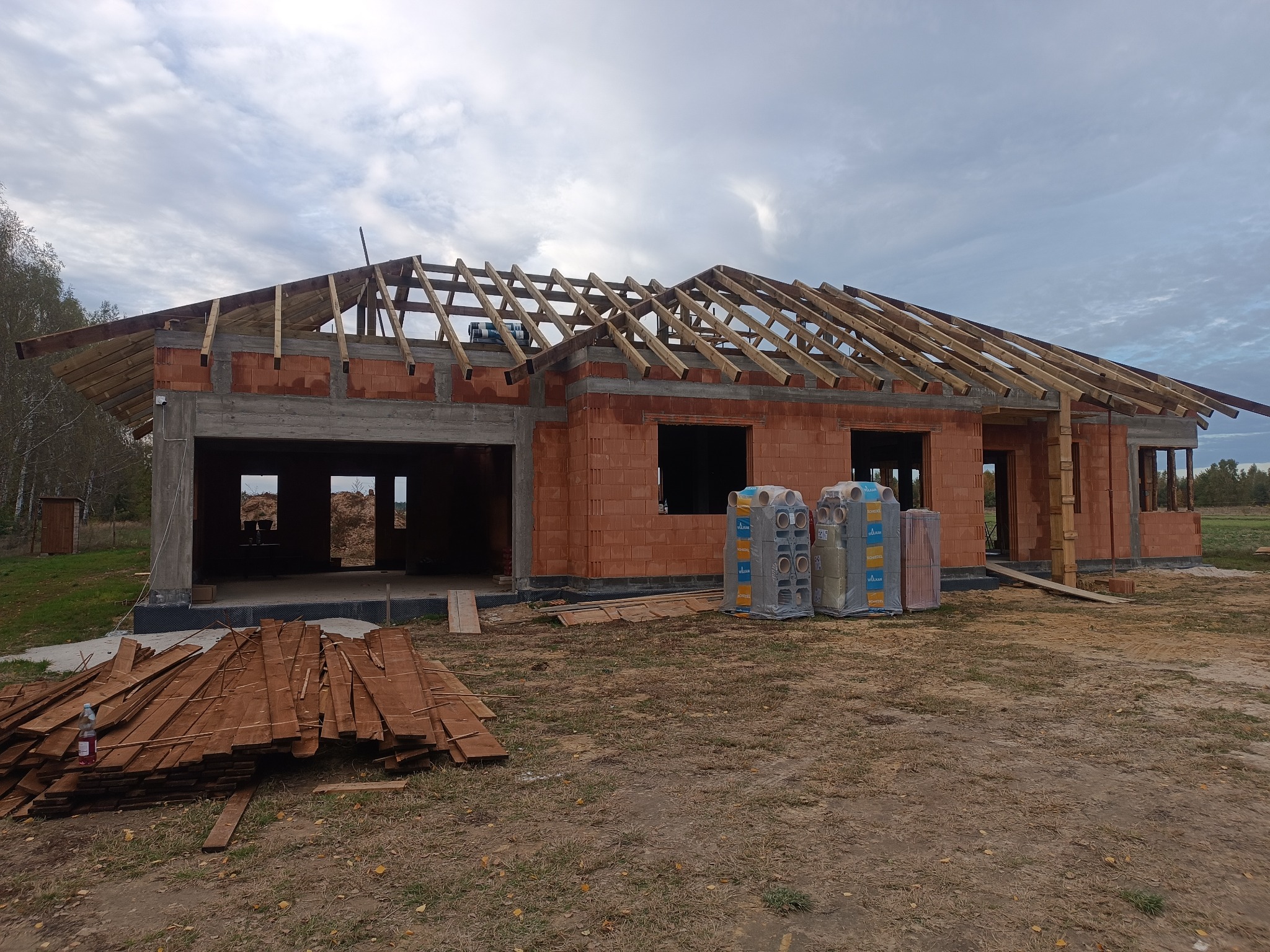 Wznoszenie domu z czerwonej cegły ceramicznej, widoczne drewniane elementy konstrukcji dachu, palety z materiałami budowlanymi oraz sterta desek na pierwszym planie.