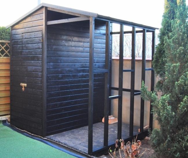 Częściowo wykończona, czarna, drewniana szopa ogrodowa z widoczną konstrukcją szkieletową bocznej ściany i zielonym trawnikiem w otoczeniu.