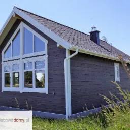 Gotowy dom z drewnianych bali pomalowany na szaro, z białymi oknami i dachem z ciemnej dachówki bitumicznej, na tle błękitnego nieba i zielonej trawy.
