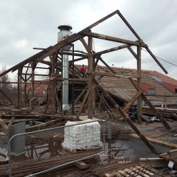 Rozbiórki i wyburzenia kompleksowo - Demontaż drewnianej konstrukcji dachu z kominem na budynku z czerwoną dachówką. Widoczne materiały budowlane i pracownik w czerwonej koszulce.