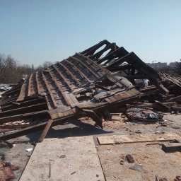 Rozbiórki i wyburzenia kompleksowo - Demontaż drewnianej konstrukcji dachu z widocznymi elementami więźby i fragmentami starej dachówki na tle błękitnego nieba i zabudowań miejskich w oddali.