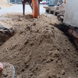 Rozbiórki i wyburzenia kompleksowo - Kupa piasku z taczką i cegłami w tle, przygotowana do prac murarskich. Widoczne narzędzia i materiały budowlane.