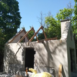 Rozbiórki i wyburzenia kompleksowo - Fragment rozbieranej, murowanej konstrukcji budynku z widocznymi elementami drewnianymi więźby dachowej na tle zieleni drzew i błękitnego nieba.