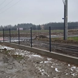 Rozbiórki i wyburzenia kompleksowo - Nowoczesne ogrodzenie panelowe z podmurówką betonową, widok z perspektywy gruntu na tle zaśnieżonego pola i pochmurnego nieba. Słup energetyczny w tle.