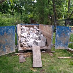 Rozbiórki i wyburzenia kompleksowo - Kontener wypełniony gruzem budowlanym na trawie, otwarte drzwi kontenera, widoczne drzewa w tle. Kontener w kolorze niebieskim, z widocznymi śladami użytkowania.