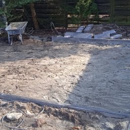 Rozbiórki i wyburzenia kompleksowo - Układanie obrzeży i kostki brukowej na przygotowanym podłożu. Widoczna taczka, drzewa w tle i częściowo ułożona kostka. Prace ziemne w toku.