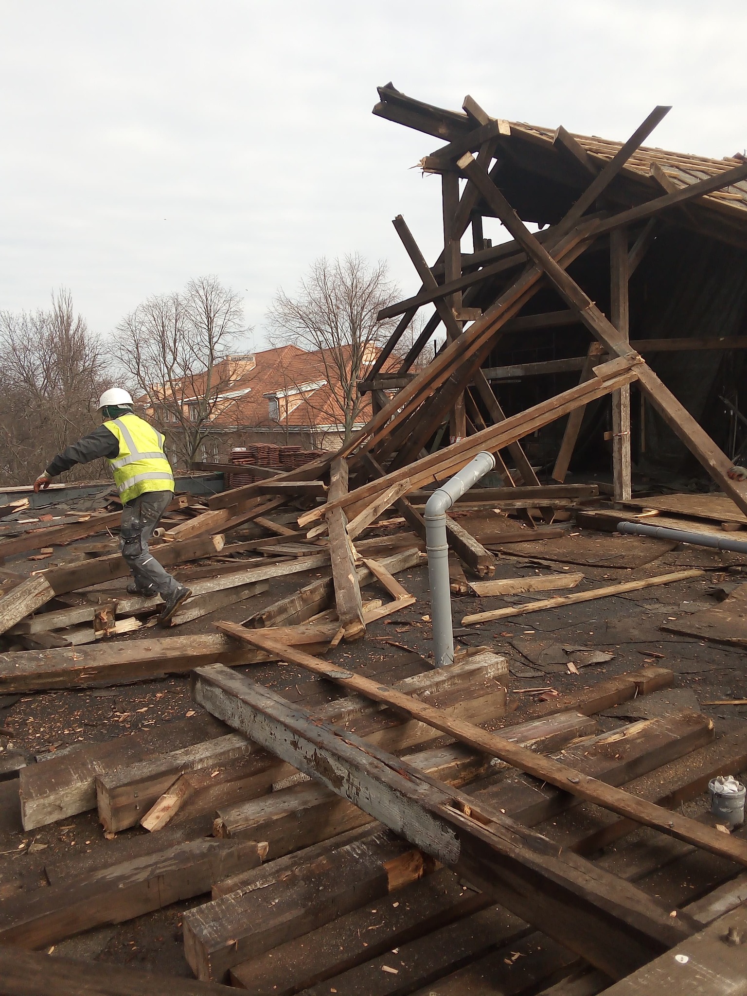 Demontaż drewnianej konstrukcji dachu przez pracownika w kasku i kamizelce odblaskowej. Widoczne elementy konstrukcyjne i rury wentylacyjne.