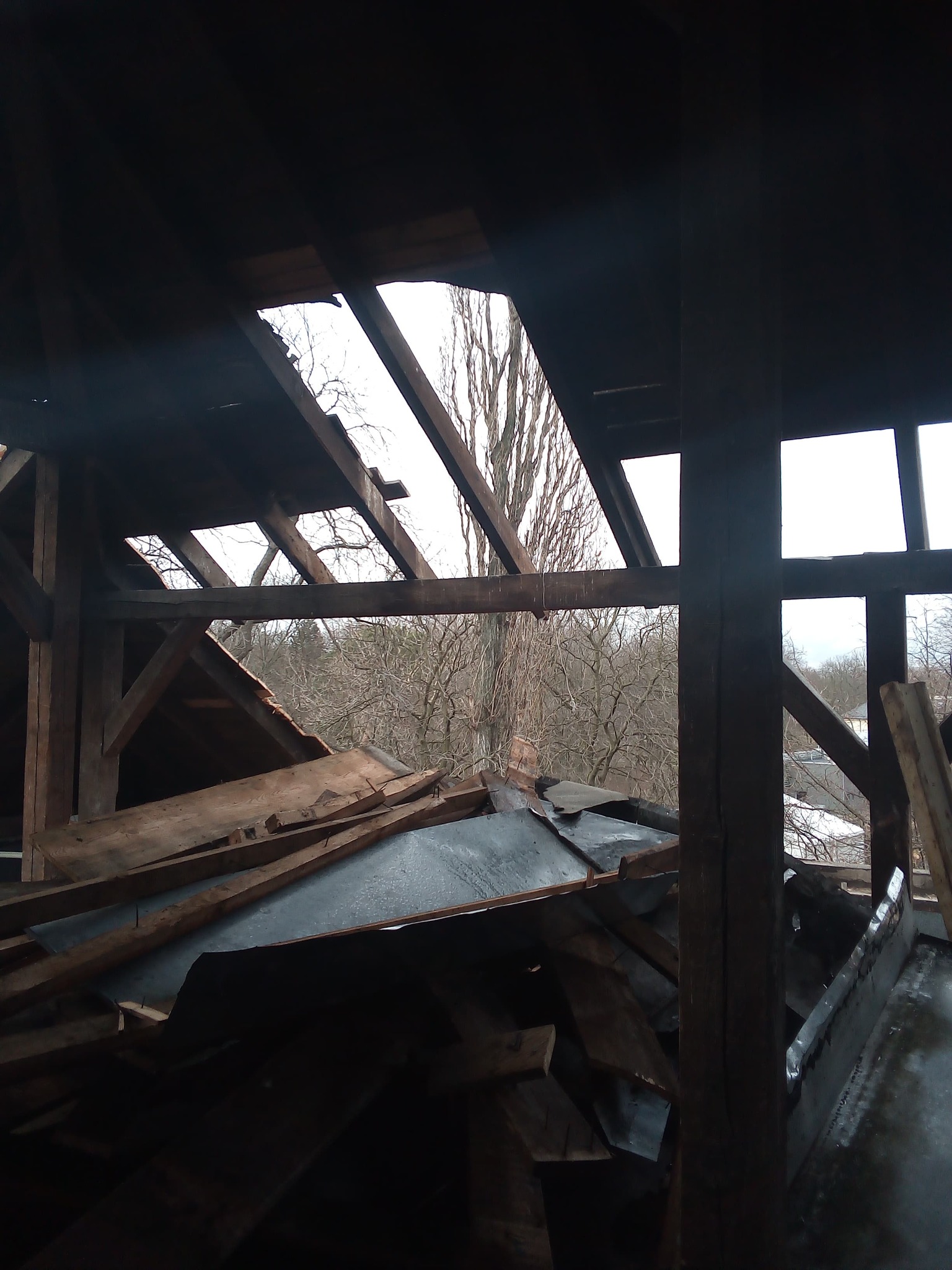 Demontaż drewnianej konstrukcji dachu z widocznymi belkami i fragmentami poszycia, z prześwitami na zewnątrz ukazującymi drzewa.