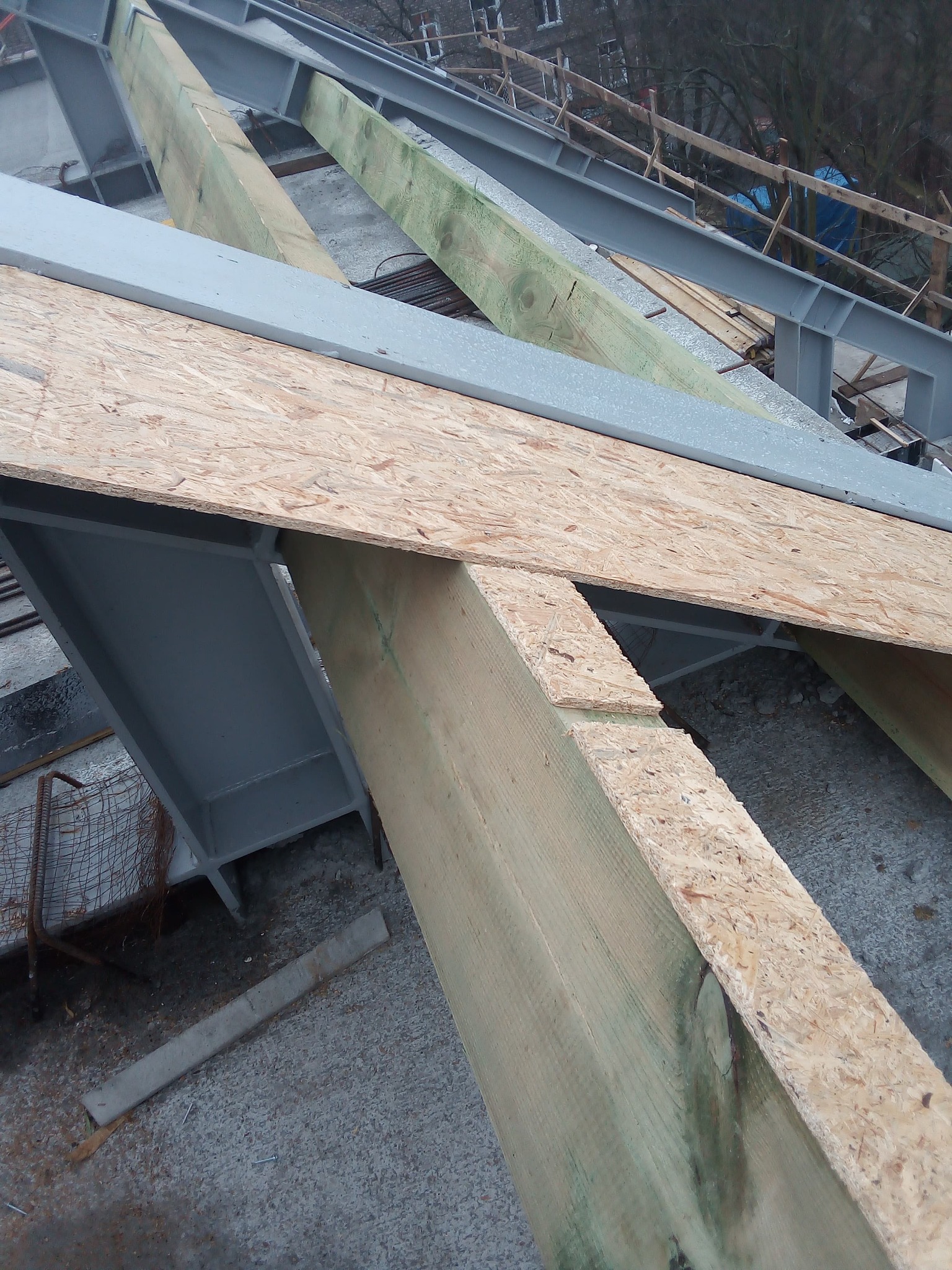 Konstrukcja stalowa z drewnianymi elementami i płytami OSB w trakcie budowy. Widoczne szare profile stalowe i surowe drewno. Ujęcie z góry, częściowo na zewnątrz.