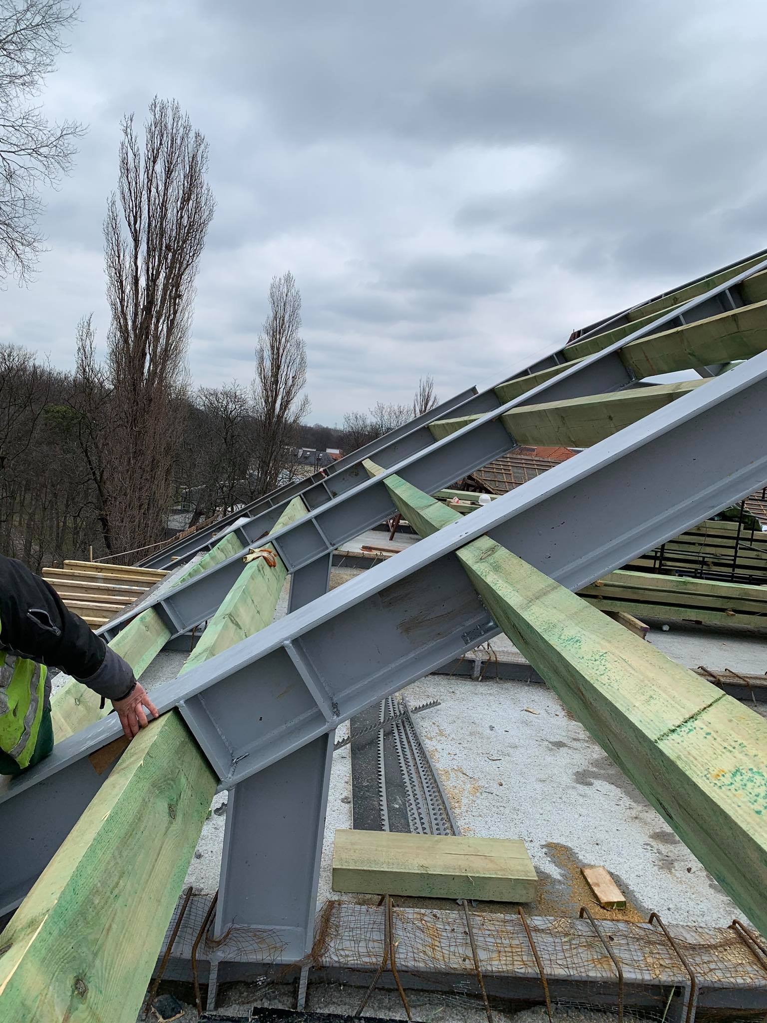 Konstrukcja dachu z belek stalowych i drewnianych łat, widok z góry, częściowo widoczna sylwetka pracownika w odblaskowej kamizelce, pochmurne niebo w tle.