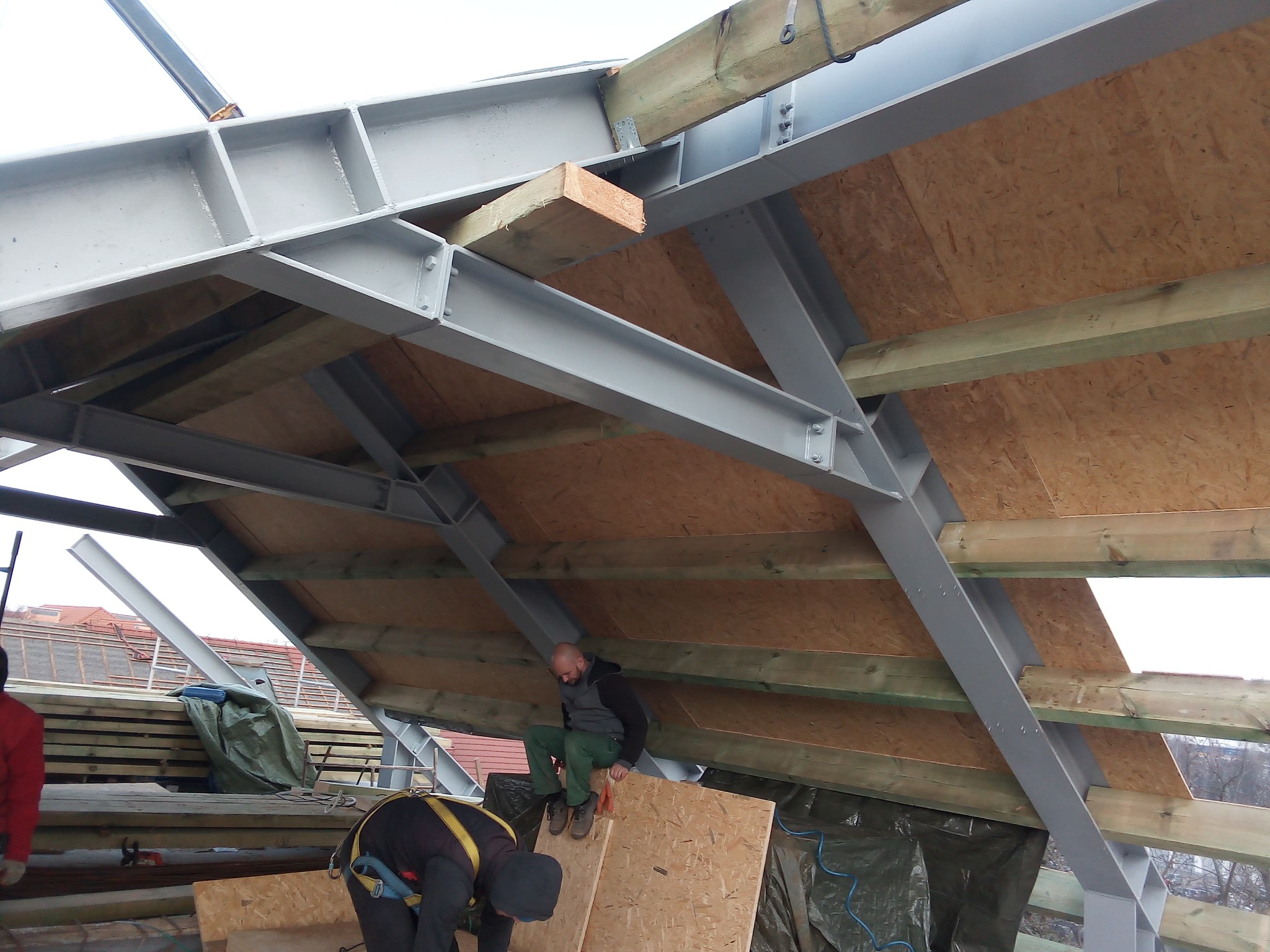 Konstrukcja dachu z belek stalowych i drewnianych łat, widoczni pracownicy w trakcie montażu, zabezpieczeni uprzężami. Prace dekarskie w toku.