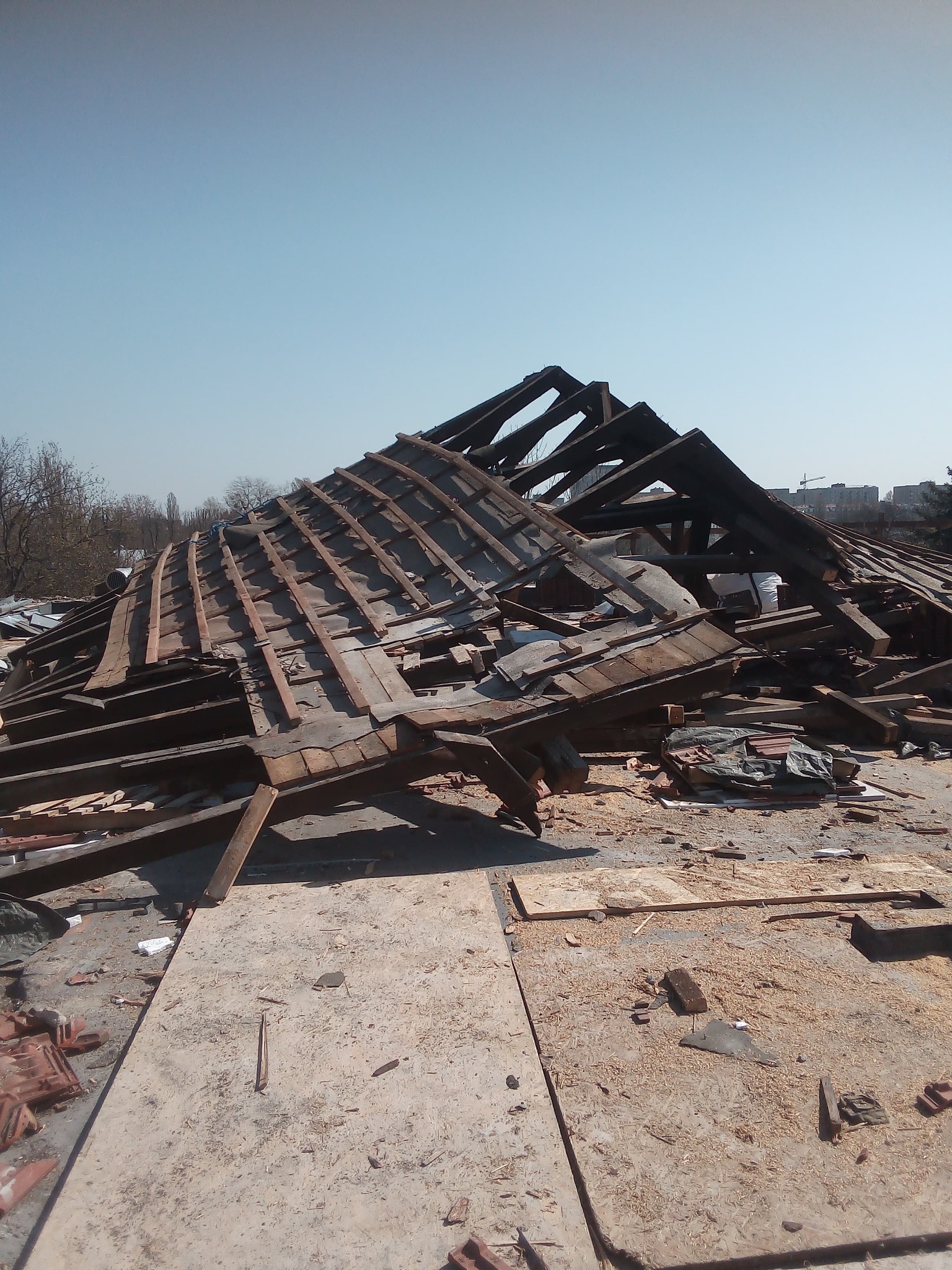 Demontaż drewnianej konstrukcji dachu z widocznymi elementami więźby i fragmentami starej dachówki na tle błękitnego nieba i zabudowań miejskich w oddali.