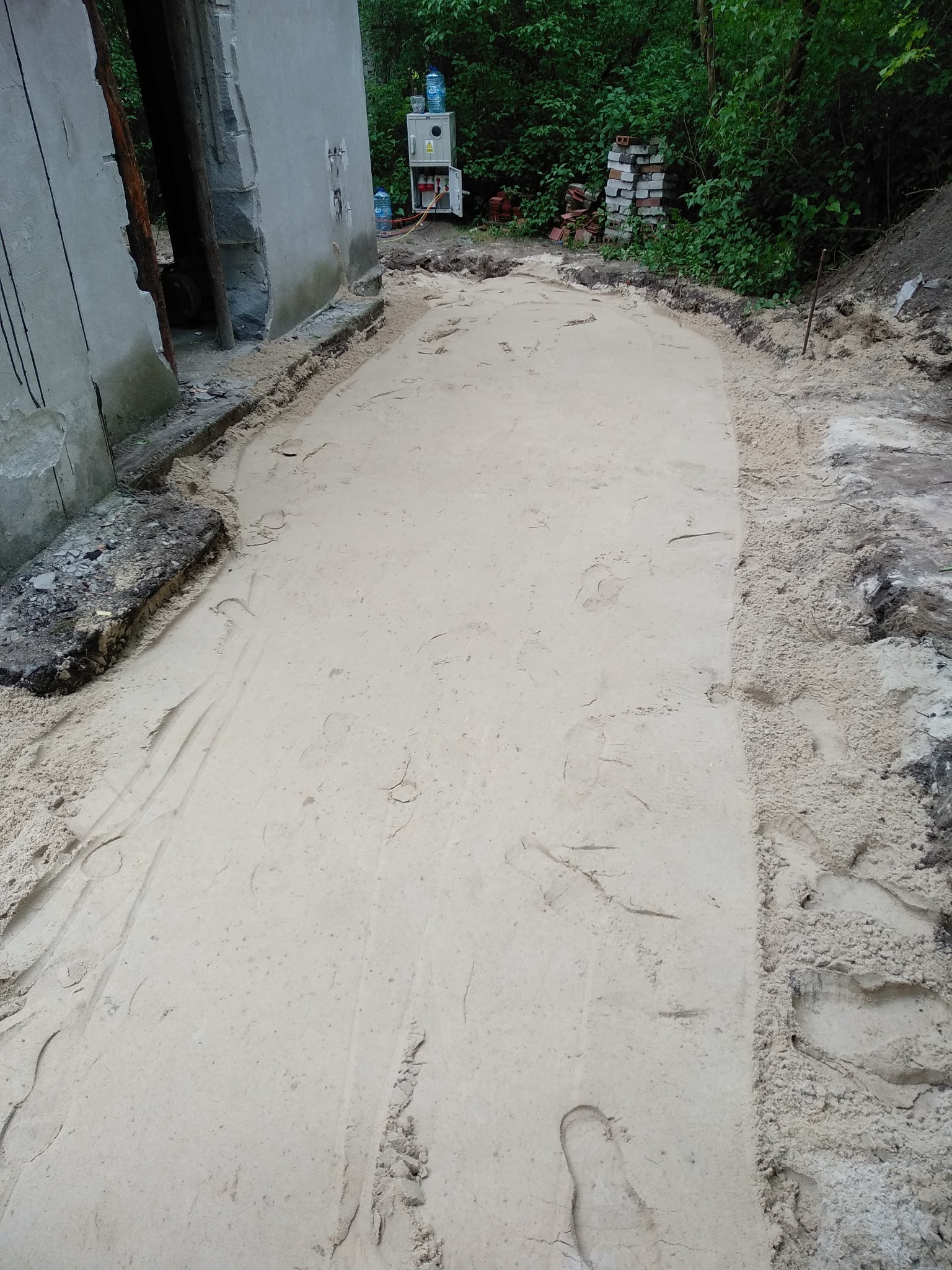 Przygotowany teren pod wylewkę betonową z widocznymi śladami stóp, obok fragment budynku w stanie surowym i sterta cegieł. Widoczne zasilanie elektryczne.