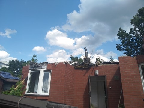 Ceglany budynek po wyburzeniach, widoczny fragment dachu z kominem, okno i drzwi. Na dachu gruz i zniszczenia. Błękitne niebo z chmurami w tle.