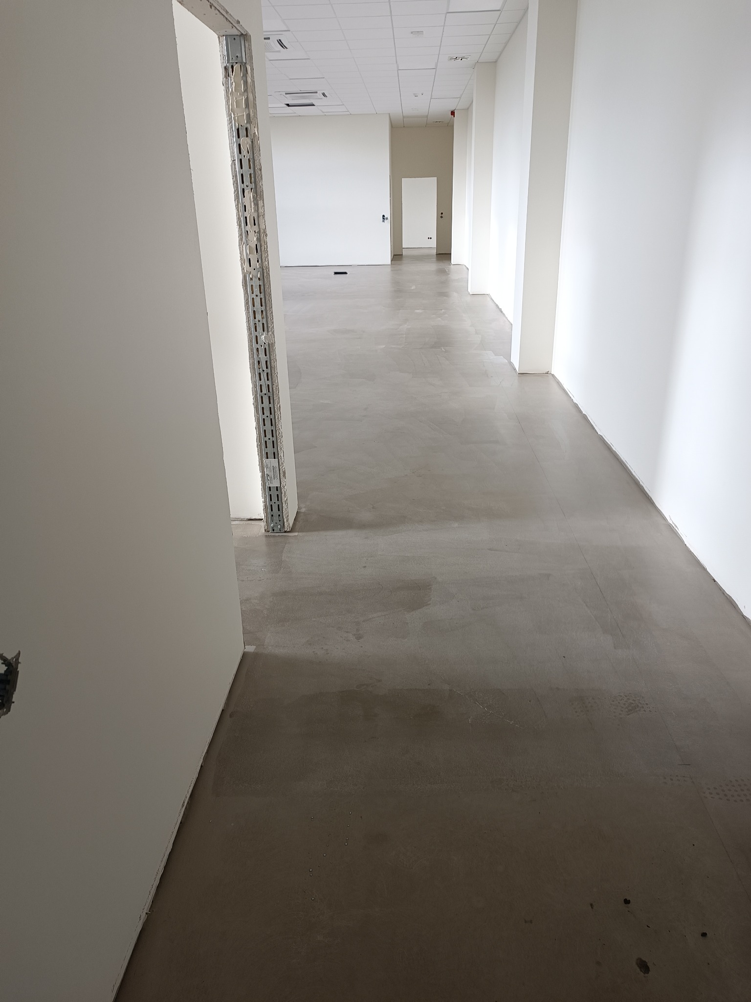 Wnętrze biura w trakcie wykończenia: surowa podłoga, białe ściany, widoczne profile montażowe. Minimalistyczny design, jasne oświetlenie, perspektywa w głąb korytarza.