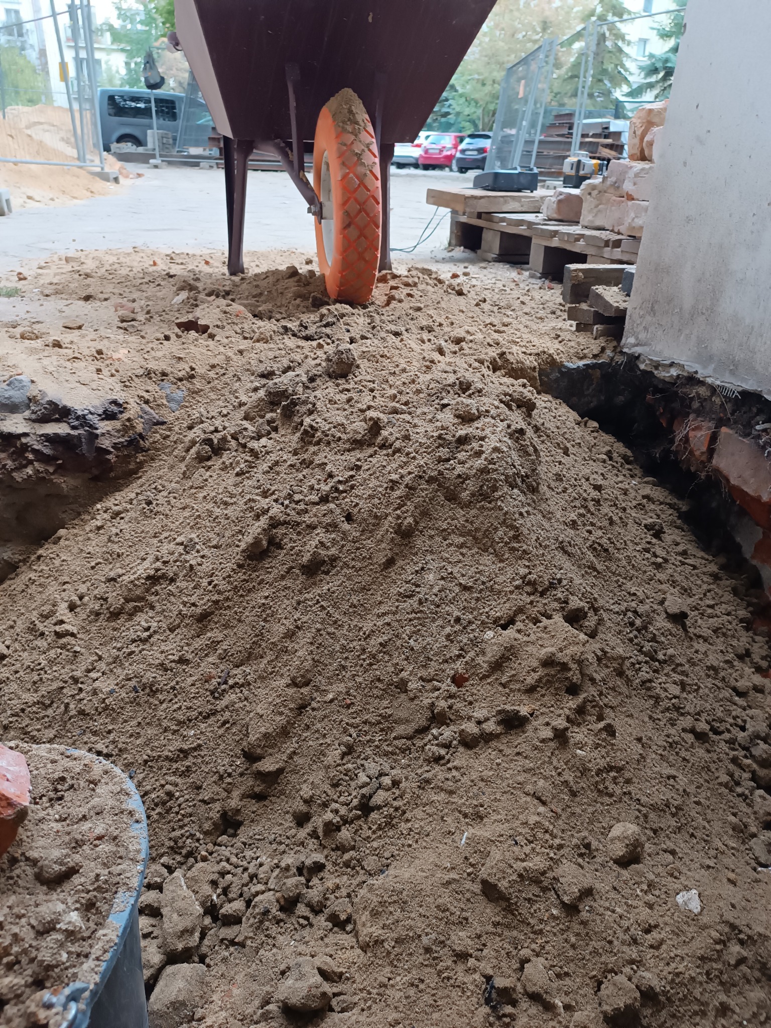 Kupa piasku z taczką i cegłami w tle, przygotowana do prac murarskich. Widoczne narzędzia i materiały budowlane.