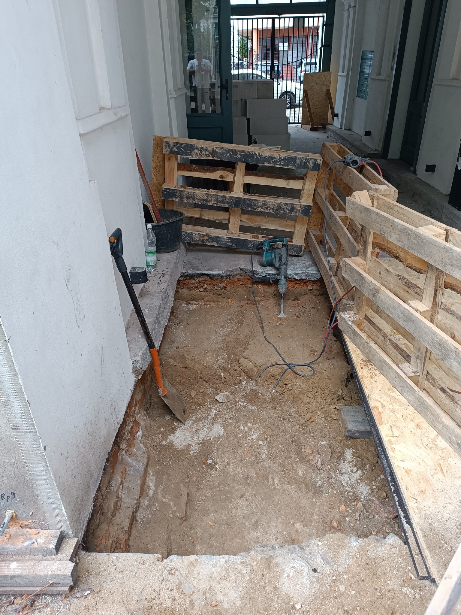 Wykop pod fundamenty z widoczną łopatą, młotem pneumatycznym i paletami. Prace ziemne w trakcie realizacji, przygotowanie do wylewki betonowej. Widoczne cegły i piasek.