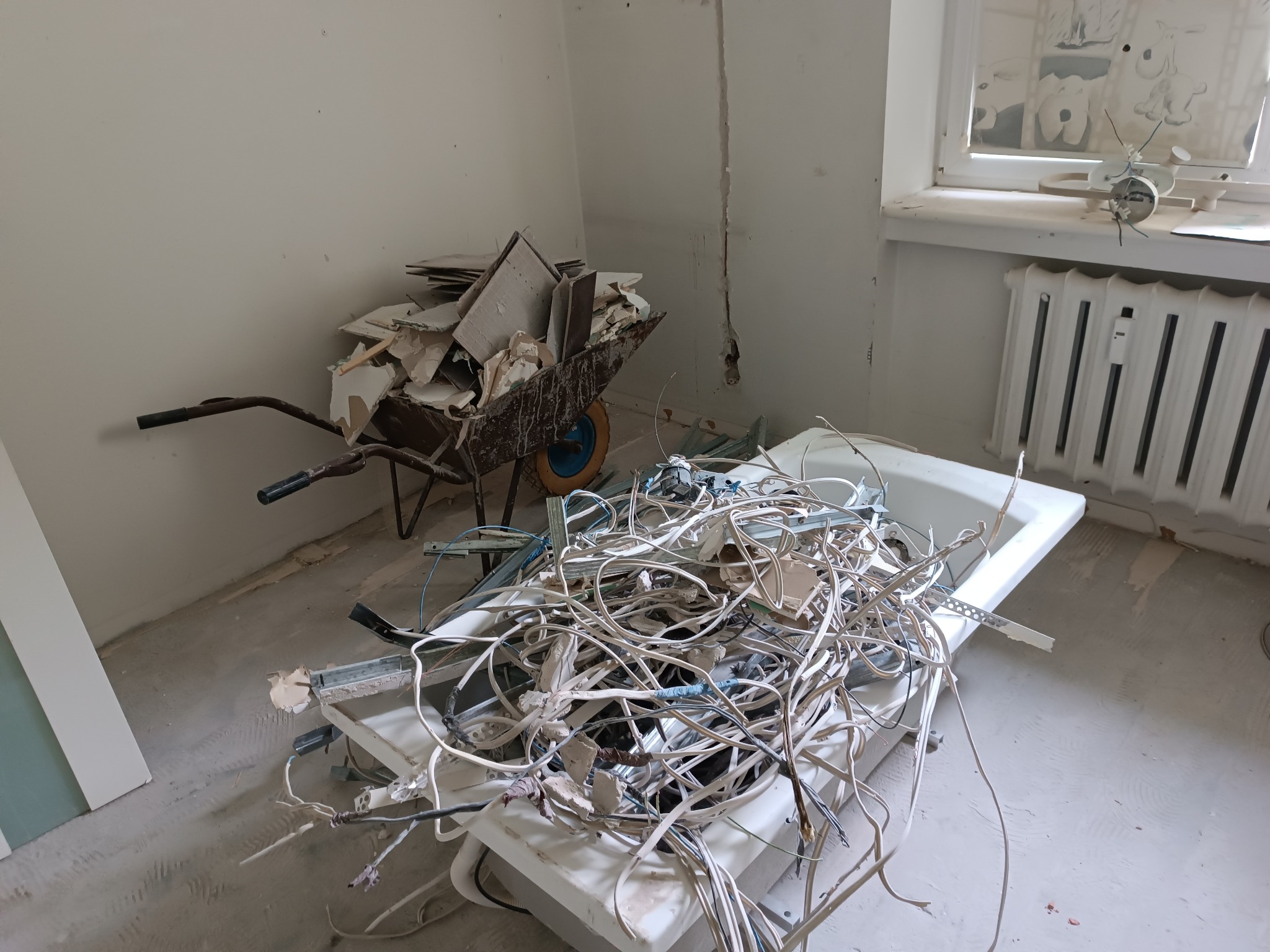 Wnętrze w trakcie remontu: gruz w taczce i stare kable w wannie. Widoczne ślady po instalacji elektrycznej na ścianie. W tle okno z zabawką.