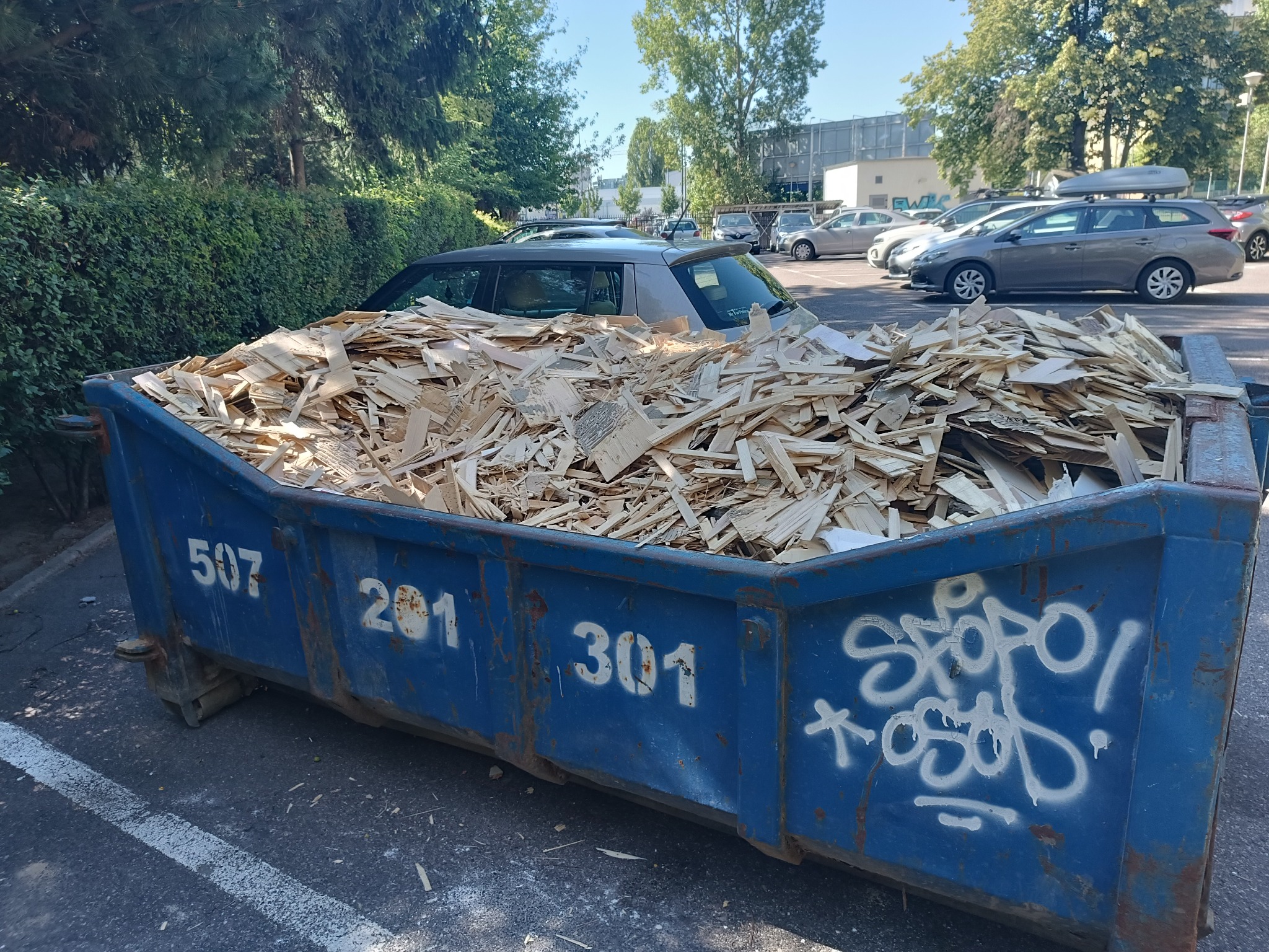 Niebieski kontener wypełniony po brzegi drewnianymi odpadami budowlanymi, z widocznymi numerami i graffiti, stojący na parkingu w słoneczny dzień.