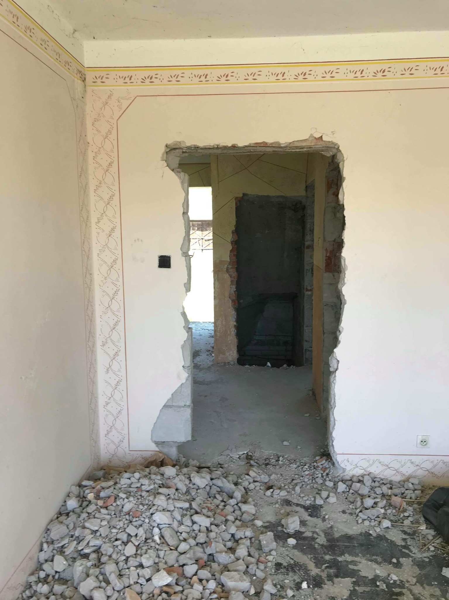 Otwór wybity w ścianie z dekoracyjną bordiurą, gruz na podłodze. Widok na sąsiednie pomieszczenie przez otwór. Etap prac remontowych.