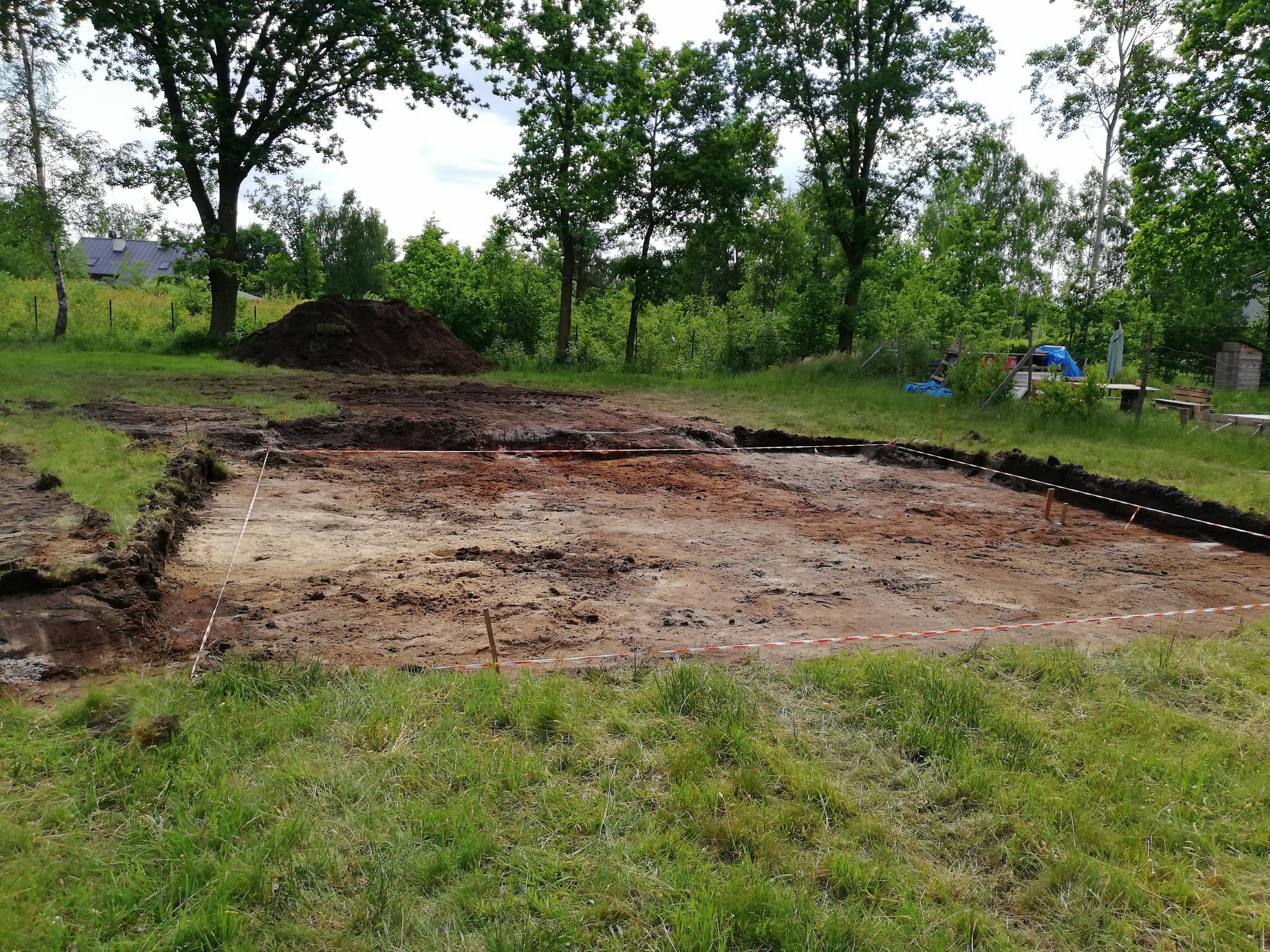 Wytyczony teren pod budowę z wykopanymi fundamentami, zabezpieczony taśmą ostrzegawczą. W tle drzewa i hałda ziemi.