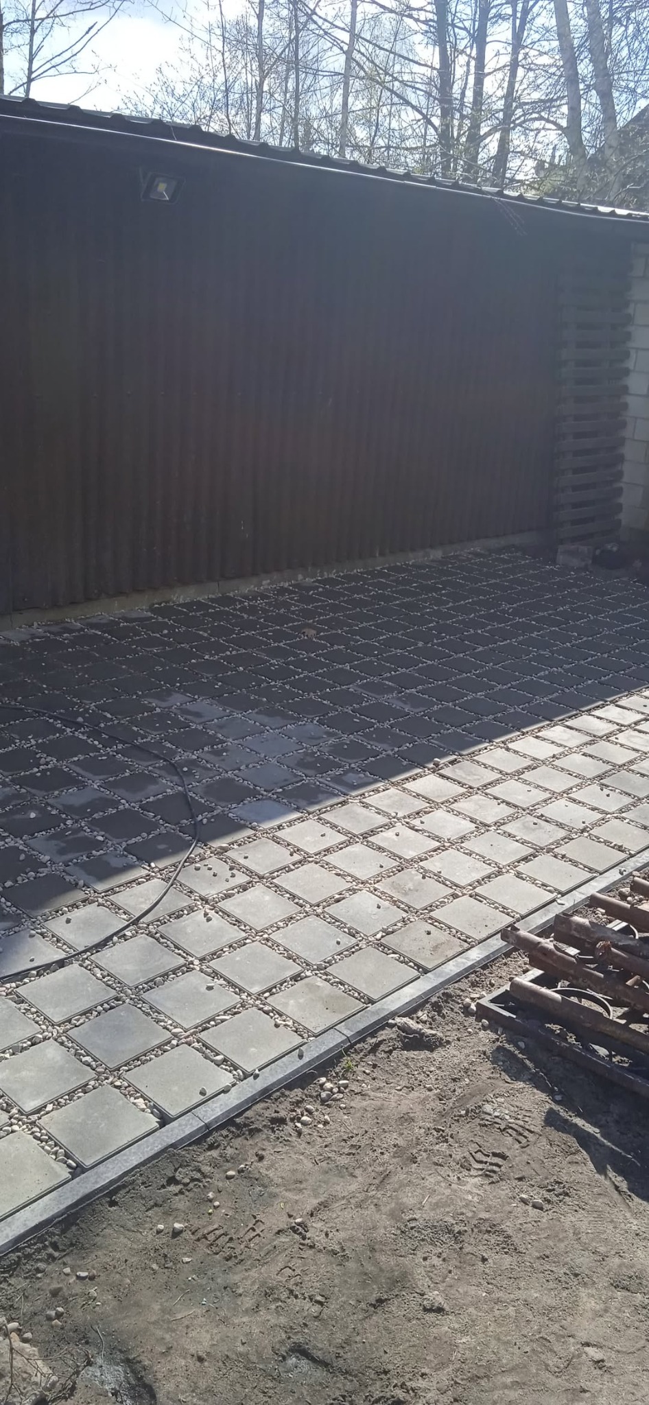 Ułożona kostka brukowa z drobnymi kamieniami w przestrzeniach, tworząca wzór szachownicy. Widoczne narzędzia i fragment budynku w tle. Słoneczny dzień.