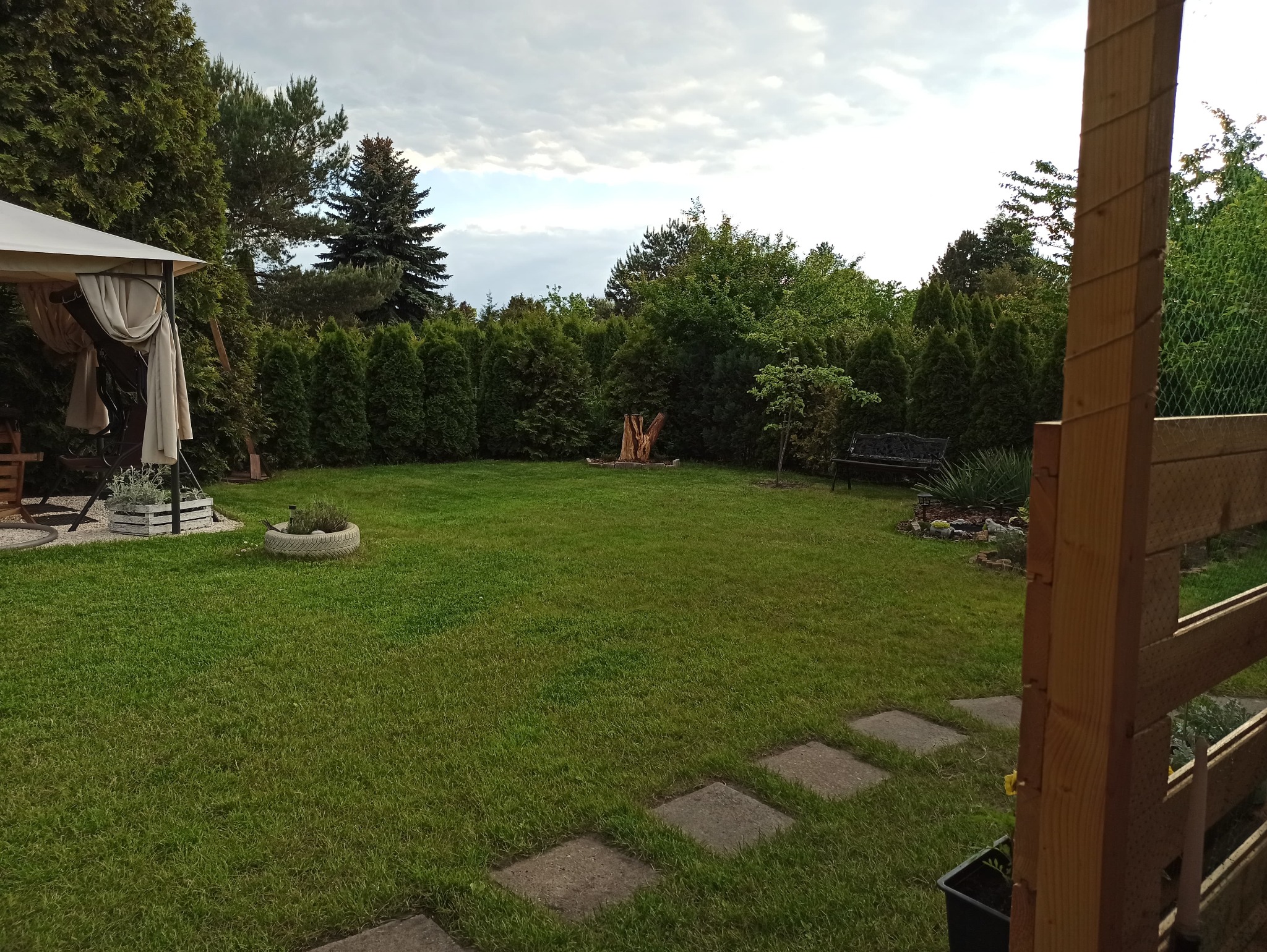 Zadbany ogród z równo przystrzyżonym trawnikiem, ozdobnymi krzewami, drzewami i elementami dekoracyjnymi, takimi jak kwietniki i ścieżka z płyt.