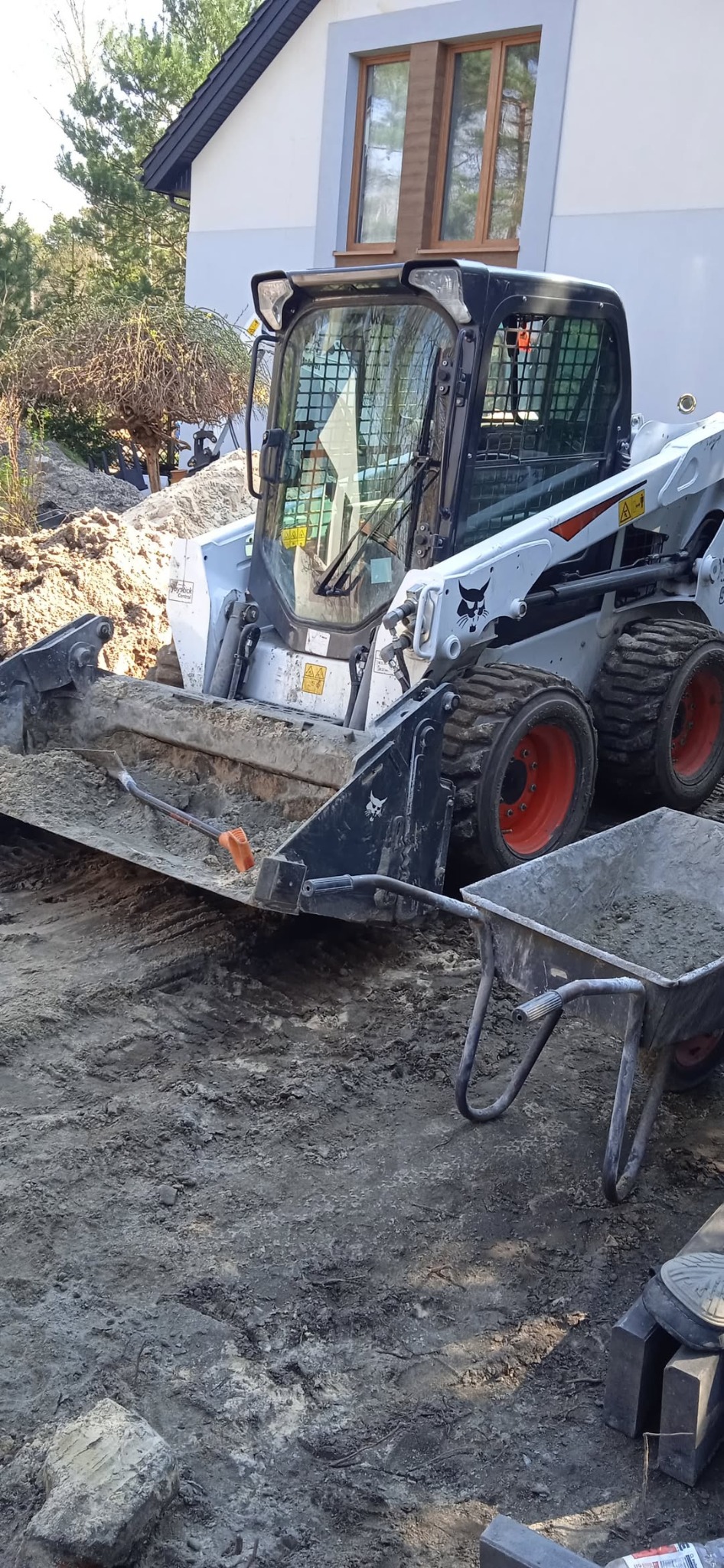 Biała miniładowarka Bobcat z łyżką i taczką na ziemi przed domem. Widoczna łopata w łyżce. Prace ziemne w toku, przygotowanie terenu pod budowę lub ogród.