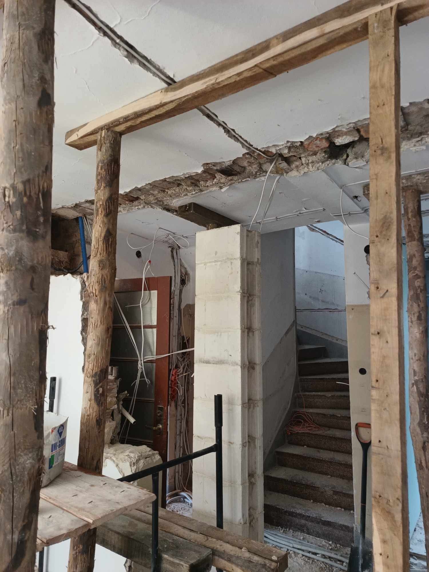Wnętrze w trakcie rozbiórki: odsłonięte przewody, fragmenty muru, drewniane podpory i schody w tle. Widoczne ślady demontażu ścian i instalacji.