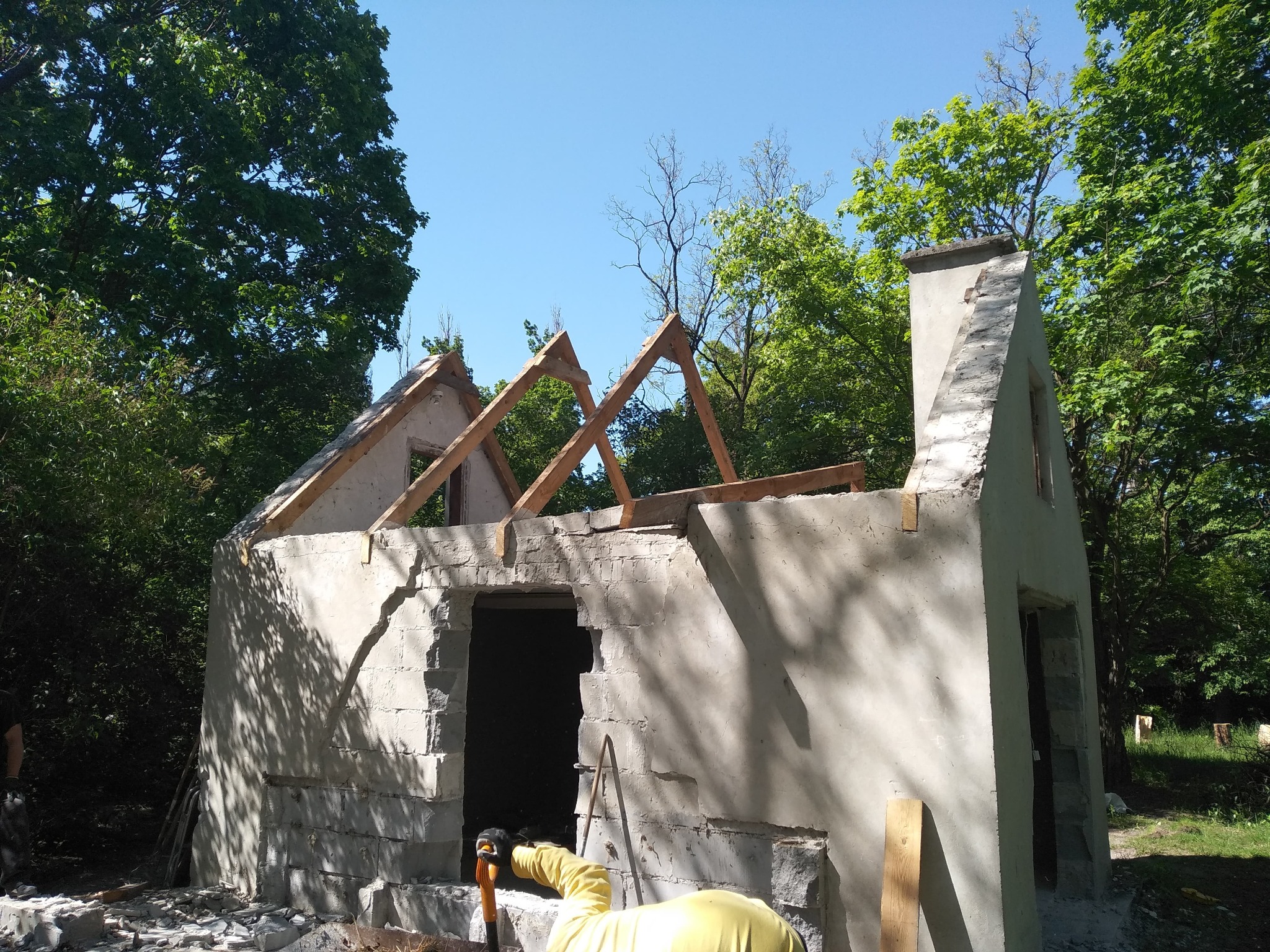 Fragment rozbieranej, murowanej konstrukcji budynku z widocznymi elementami drewnianymi więźby dachowej na tle zieleni drzew i błękitnego nieba.