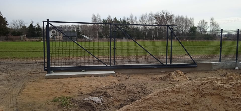 Nowoczesna, przesuwna brama panelowa w kolorze grafitowym, zamontowana na betonowej podstawie. W tle zielone pole i drzewa. Widoczne ślady prac ziemnych.