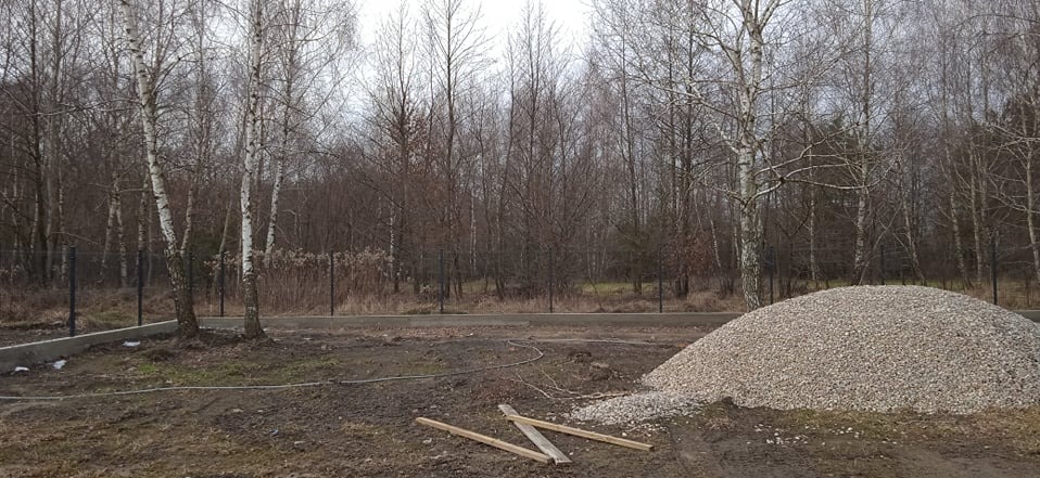 Fundament pod ogrodzenie na tle lasu brzozowego. Na pierwszym planie pryzma żwiru i drewniane elementy szalunku. Widoczne ślady prac ziemnych.