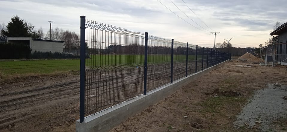 Długa, metalowa siatka ogrodzeniowa na betonowej podmurówce, oddzielająca teren budowy od zielonego pola. Słupki ogrodzenia równomiernie rozstawione.