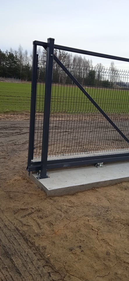 Fragment przesuwnej bramy panelowej z metalową konstrukcją, osadzonej na betonowej podstawie. W tle pole i drzewa.