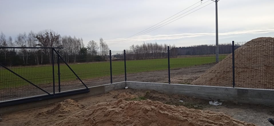 Nowoczesne ogrodzenie panelowe z bramą przesuwną na betonowej podmurówce, widok na zielone pole i las w tle, pochmurne niebo.