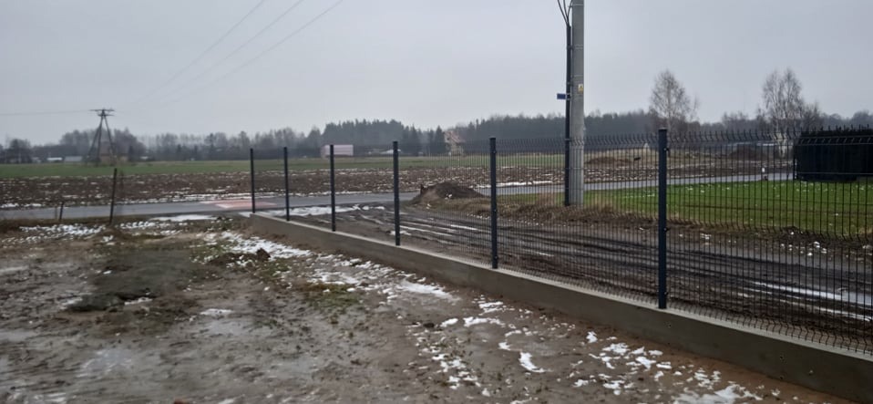 Nowoczesne ogrodzenie panelowe z podmurówką betonową, widok z perspektywy gruntu na tle zaśnieżonego pola i pochmurnego nieba. Słup energetyczny w tle.