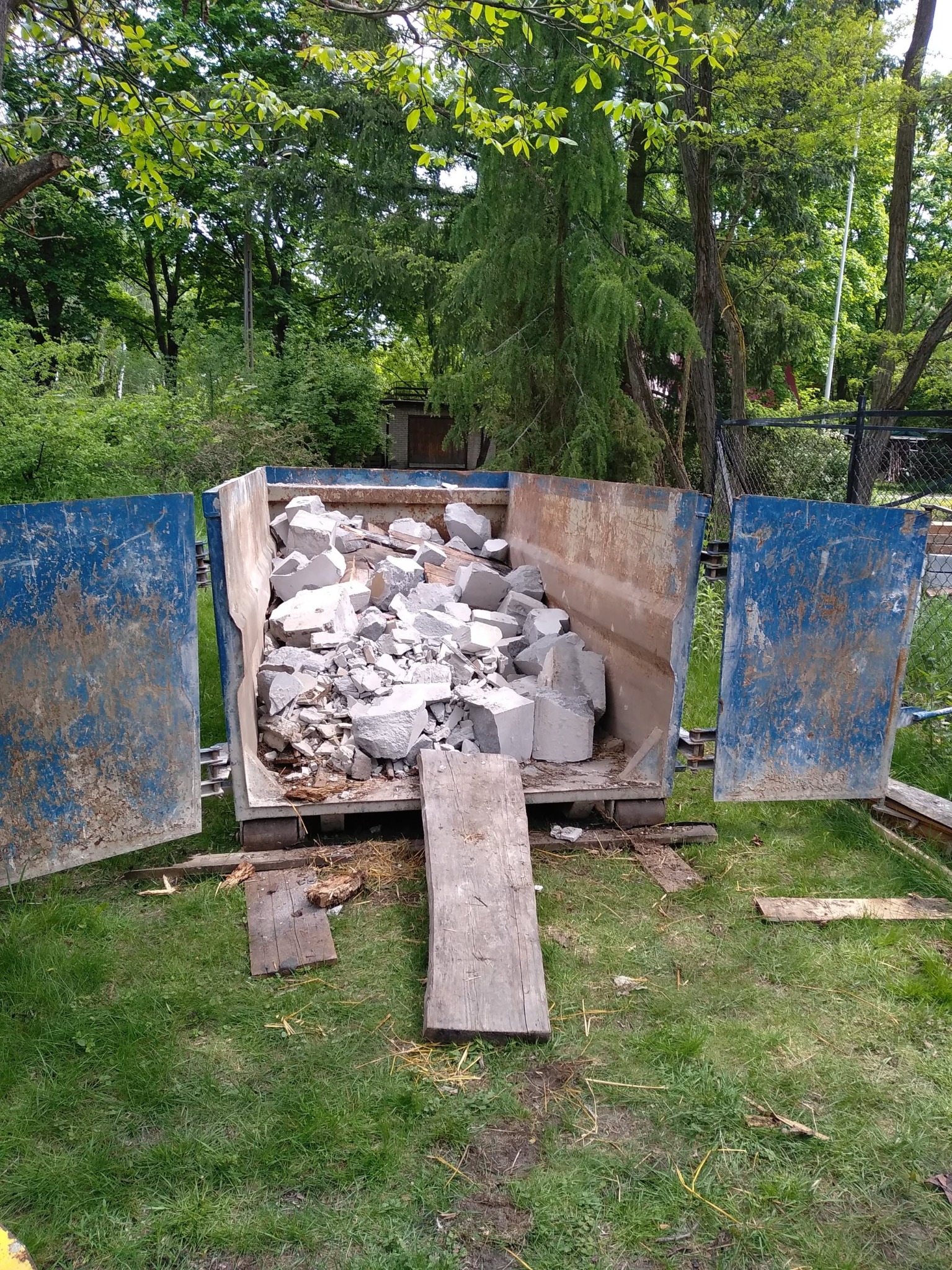 Kontener wypełniony gruzem budowlanym na trawie, otwarte drzwi kontenera, widoczne drzewa w tle. Kontener w kolorze niebieskim, z widocznymi śladami użytkowania.