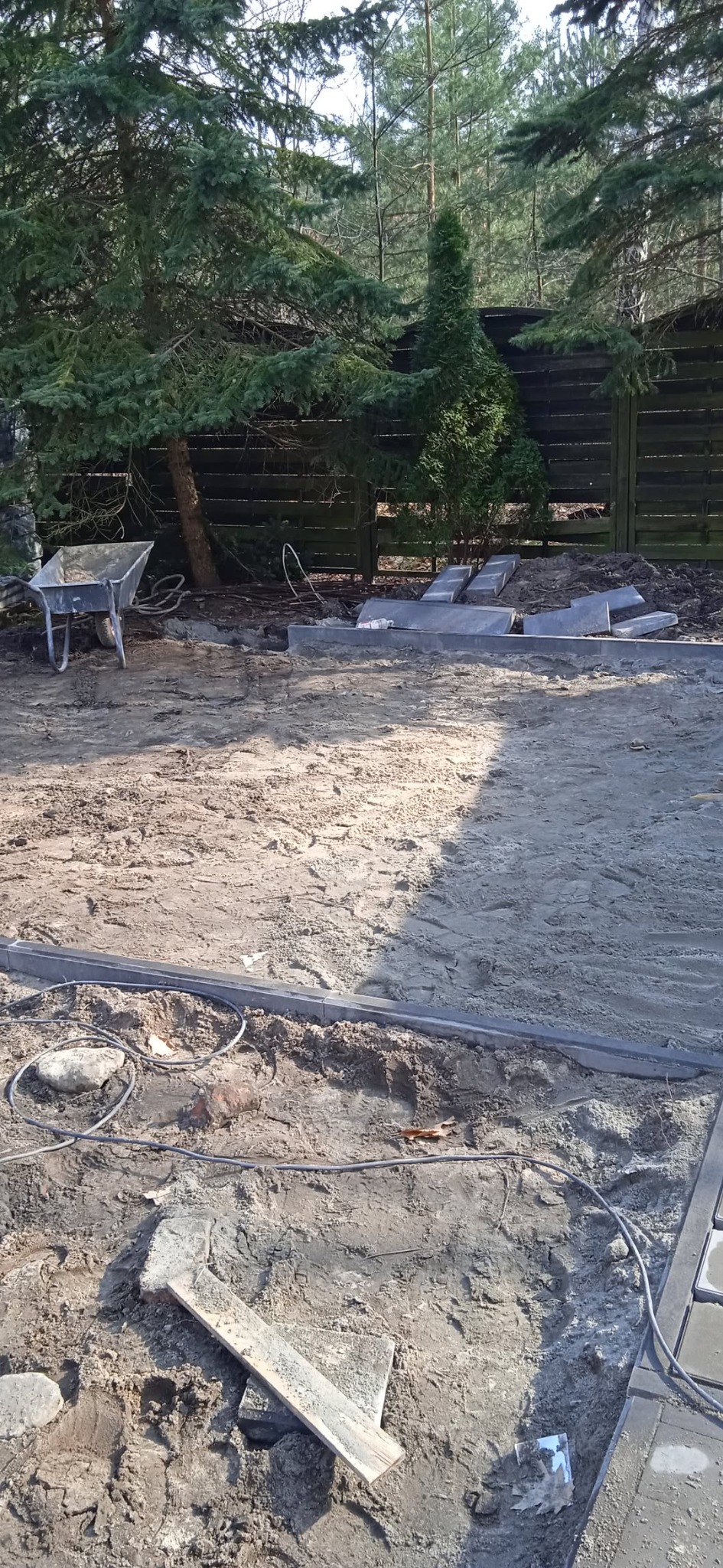 Układanie obrzeży i kostki brukowej na przygotowanym podłożu. Widoczna taczka, drzewa w tle i częściowo ułożona kostka. Prace ziemne w toku.