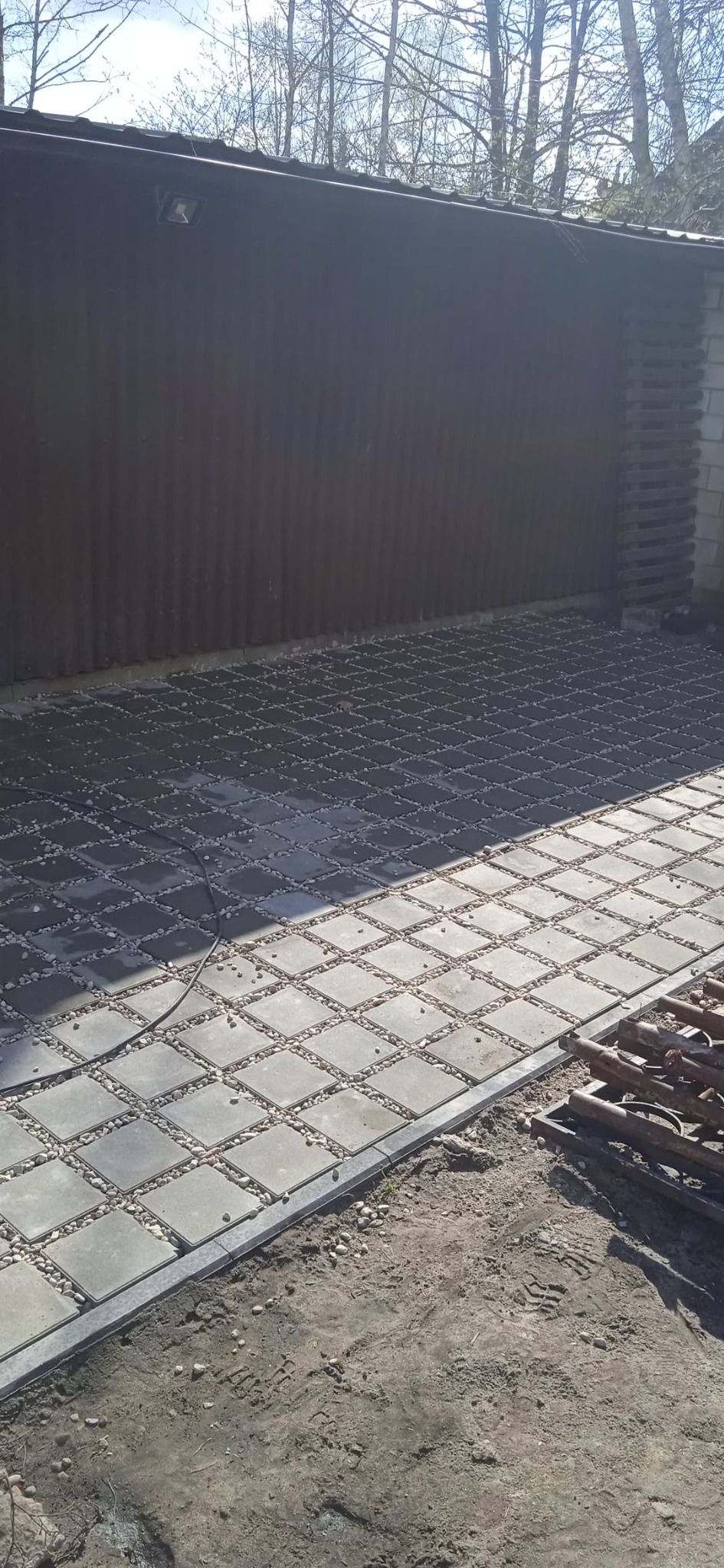Ułożona kostka brukowa z drobnymi kamieniami w przestrzeniach, tworząca wzór szachownicy. Widoczne narzędzia i fragment budynku w tle. Słoneczny dzień.