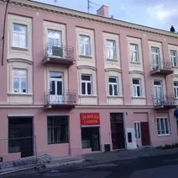 Odnowiona, trzypiętrowa kamienica w odcieniach różu i beżu, z balkonami z kutą balustradą, widoczna z poziomu ulicy, z elementami rusztowania i reklamami sklepów na parterze.