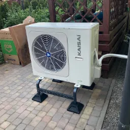 Biała jednostka zewnętrzna pompy ciepła Kaisai zamontowana na czarnym metalowym stelażu, obok kartonowe opakowanie, na tle drewnianej altany i zieleni.