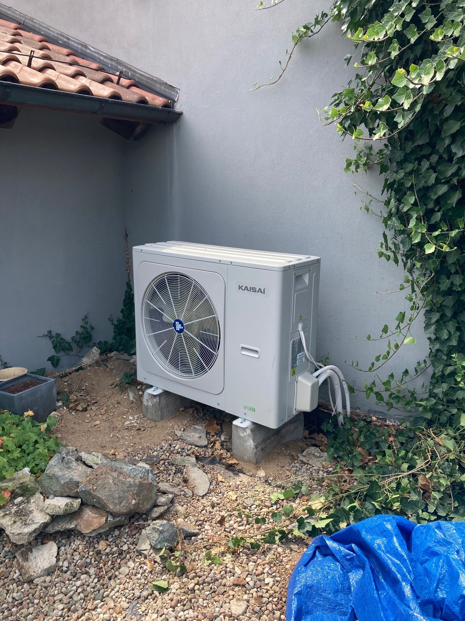 Zewnętrzna jednostka klimatyzacji Kaisai zamontowana na betonowych blokach przy szarej ścianie budynku, obok ściany porośniętej bluszczem, z niebieską plandeką w tle.