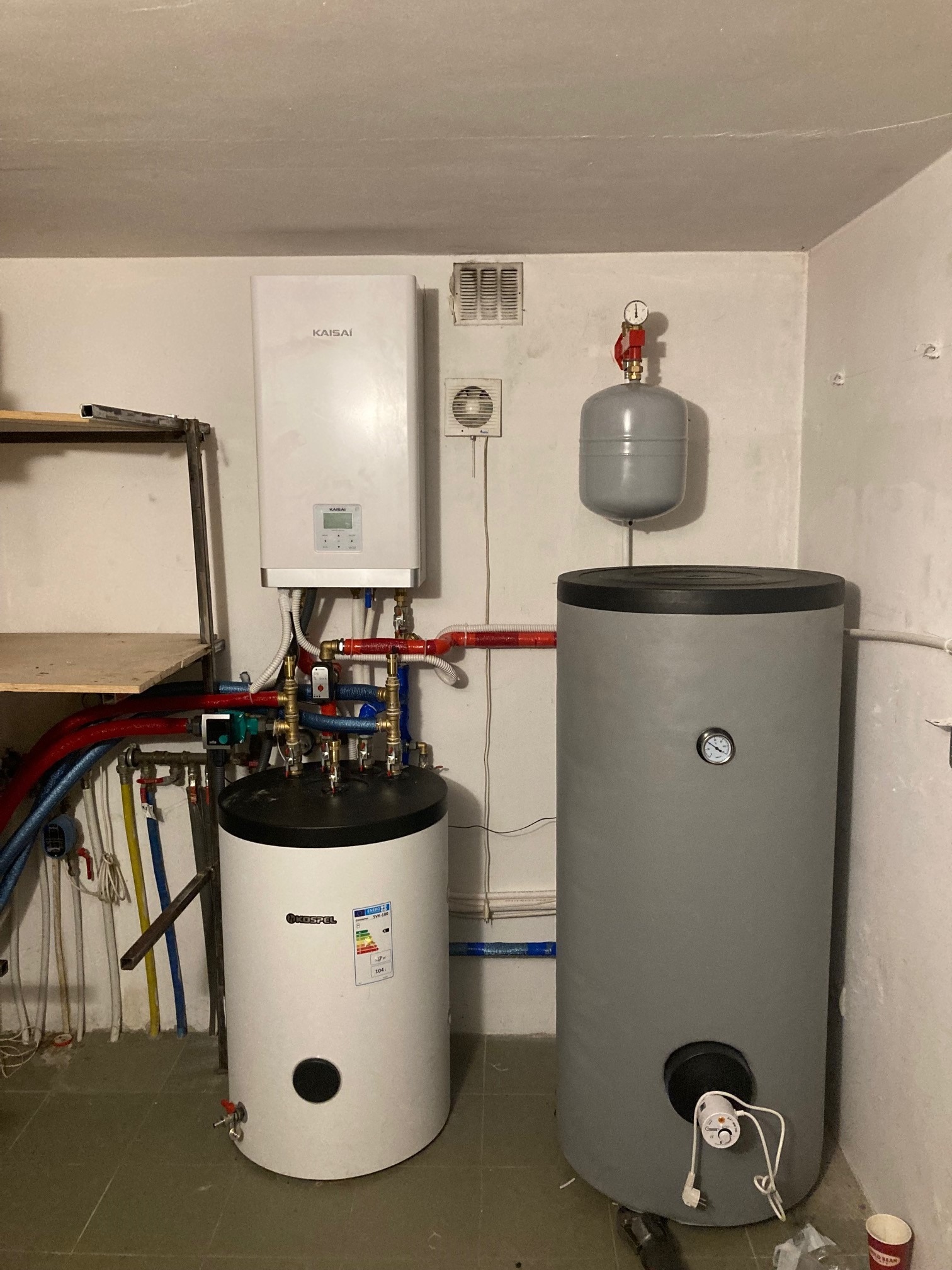 Zestaw urządzeń grzewczych w piwnicy: biała pompa ciepła Kaisai, biały zbiornik Kospel i szary zbiornik buforowy, połączone rurami z licznymi zaworami, na tle surowych, białych ścian i betonowej...