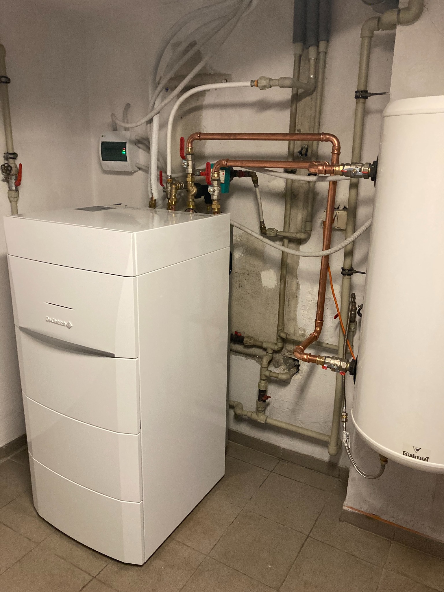 Biały piec grzewczy De Dietrich z podłączonymi miedzianymi rurami, widoczne zawory i biały zasobnik Galmet w kotłowni z szarymi płytkami podłogowymi.