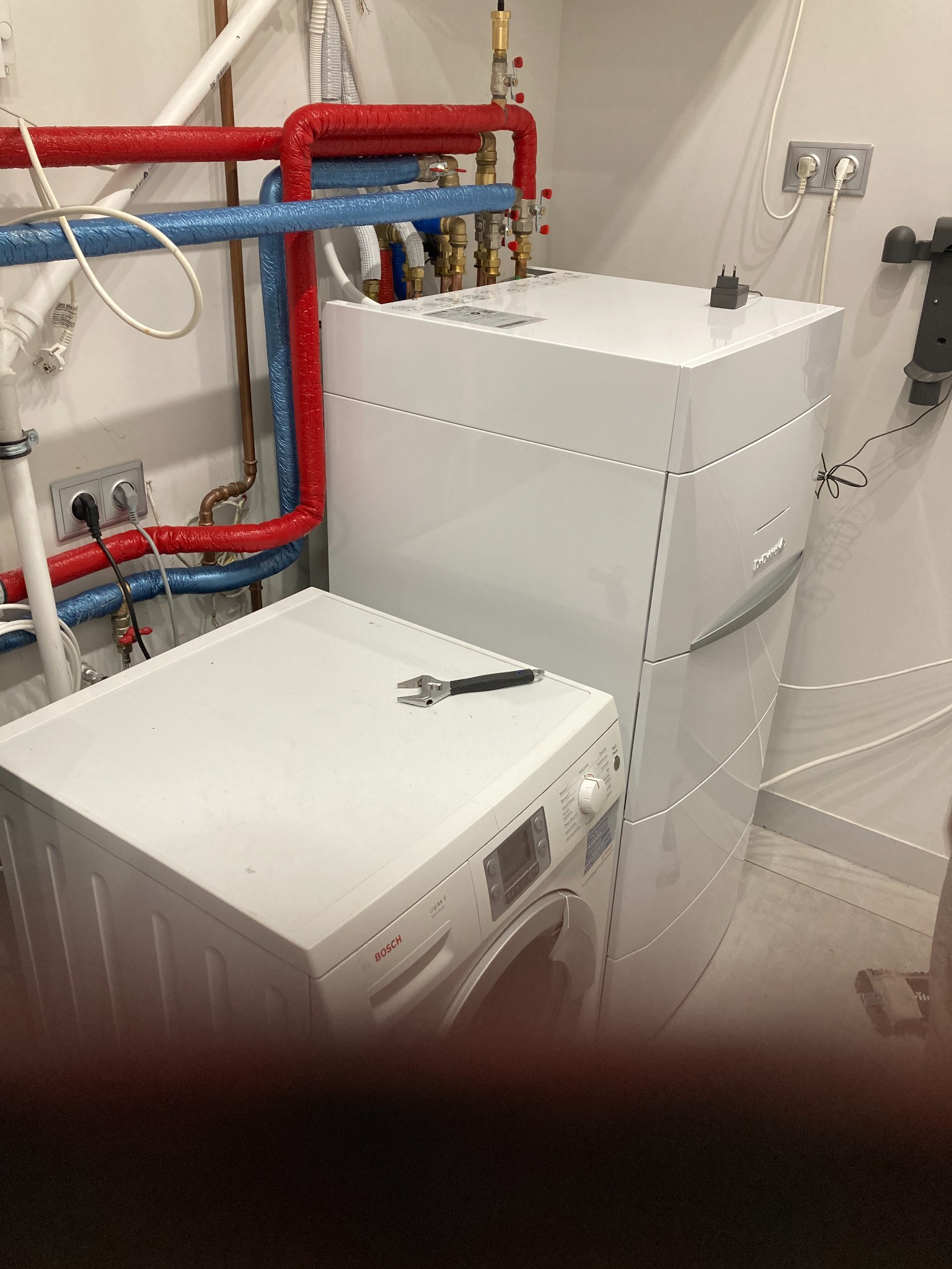 Biała pompa ciepła z widocznymi rurami w izolacji w kolorze czerwonym i niebieskim, zamontowana obok pralki w Lubiczu. Na pralce leży klucz francuski.