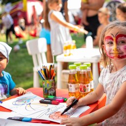 ArtGate - Dwoje dzieci siedzi przy pomarańczowym stole, rysując kredkami i flamastrami na papierze. Dziewczynka ma pomalowaną twarz w motyla.